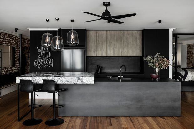 A dramatic open-plan kitchen with a large dark concrete-look island, marble-look benchtop extension and black bar stools. Black cabinetry, a timber-toned overhead cabinet, integrated appliances, a dark splashback and pendant lights create a moody industrial interior. Exposed brick walls, timber flooring, a ceiling fan and a bedroom visible through the opening add character.