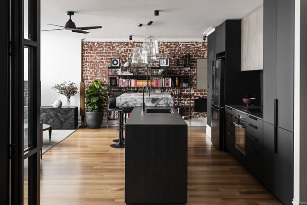 A contemporary open-plan kitchen and living area with a large dark island, black built-in cabinetry, integrated appliances, timber flooring, and a striking exposed red-brick feature wall. Open shelving displays books and decor, while black-framed glazing, a ceiling fan, and modern pendant lighting contribute to an urban industrial character.