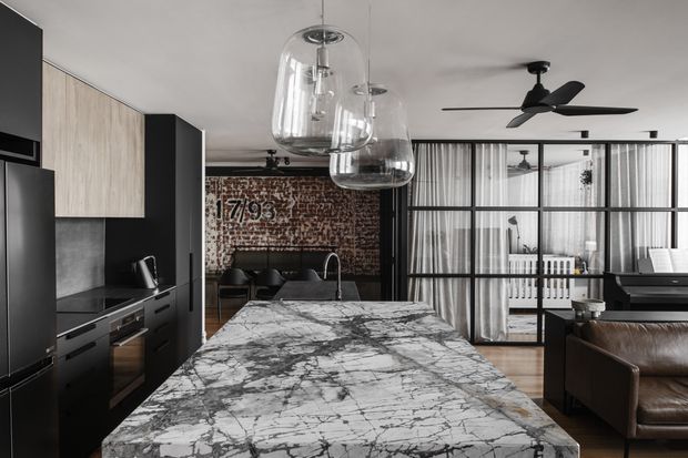 A sophisticated open-plan kitchen and living area dominated by a large veined stone island benchtop. Matte black cabinetry contrasts with pale timber-look cabinet fronts, while clear glass pendant lights hang above the island. Behind the kitchen is a dining area with a dark table and chairs, an exposed brick feature wall, and industrial-style black-framed glazing with sheer curtains. A leather sofa and ceiling fan are visible in the adjoining living area.