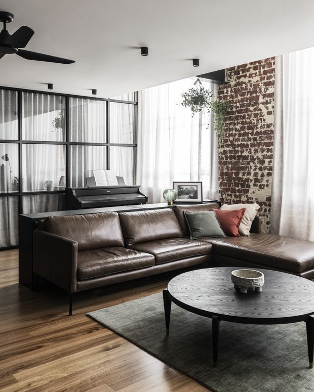 A refined industrial-style living room with a large brown leather sectional sofa, round dark coffee table, and muted area rug. The room features exposed red brick, black-framed glass partitioning, sheer full-height curtains, timber flooring, a piano, and minimalist black ceiling fixtures and fan. Abundant natural light creates a warm, sophisticated atmosphere.