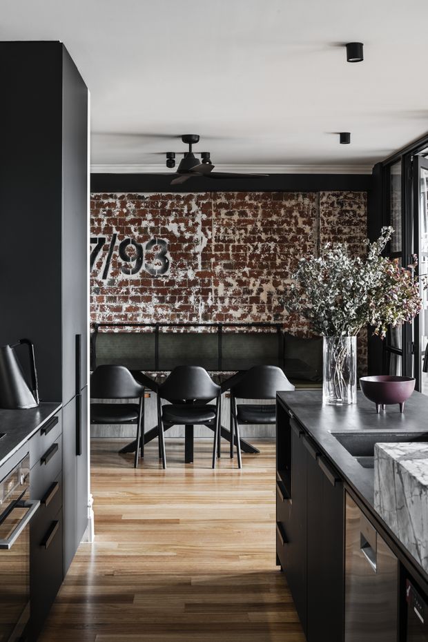 A dark, sophisticated open-plan kitchen and dining area. Matte-black cabinetry and island frame a warm timber floor, while a weathered exposed brick feature wall anchors a built-in bench and four black dining chairs. Black-framed glazing, a sculptural ceiling fan, recessed spotlights, and a marble-look island benchtop complete the industrial-contemporary interior.