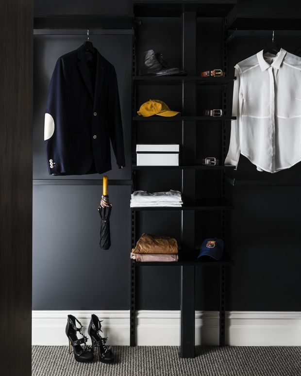 A dark, carefully organised wardrobe and dressing storage system with black shelving, hanging rails, and compartments. A navy blazer and white shirt hang on either side, while shoes, belts, hats, folded clothing, boxes, and an umbrella are displayed throughout. White skirting boards and grey carpet contrast with the predominantly black built-in cabinetry, creating a refined, moody dressing area.