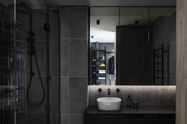 Dark, contemporary bathroom with a walk-in glass shower, black fixtures, large grey wall tiles, a floating dark vanity with a white vessel basin, and a wide mirror with concealed strip lighting. A wardrobe or dressing area is visible through the opening beyond the bathroom.