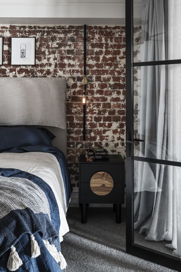 A close-up of a stylish bedroom corner featuring a bed with layered navy, white and grey bedding, a grey upholstered headboard, and a distressed exposed brick feature wall. A black metal-framed glass door with sheer curtains stands beside a compact black bedside table with a round timber inset. A slim hanging pendant light and framed artwork add detail. The room has a moody industrial character softened by textiles and warm timber accents.