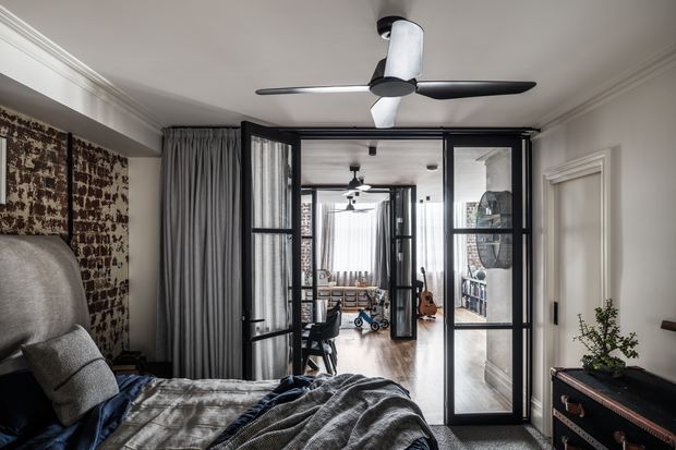 A refined bedroom viewed toward a black-framed glazed partition and hinged glass door opening onto a compact home office or study area. The room features a large upholstered bed with layered grey bedding, full-height grey curtains, patterned wall covering behind the bed, white decorative ceiling cornices and wall mouldings, a contemporary ceiling fan, and a dark timber chest with a potted plant. The glazed partition creates visual connections and reflections into the adjoining workspace.