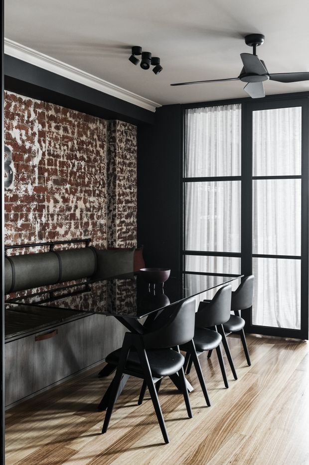 A moody dining area with a long black dining table and four upholstered dining chairs, positioned beside tall black-framed glazed doors with sheer curtains. The room features a dramatic exposed brick feature wall, dark painted walls and cabinetry, light timber flooring, white decorative ceiling cornices, and a modern black ceiling fan and track lighting.