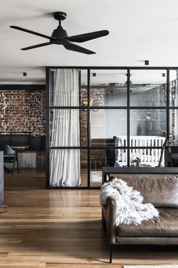 A contemporary industrial-style living room with warm timber flooring, exposed brick walls, black-framed steel-and-glass partitions, and a brown leather sofa draped with a white textured throw. A large black ceiling fan and recessed spotlights punctuate the white ceiling. Behind the glazed partition, a nursery with a white cot is visible, while dark built-in seating and cabinetry occupy the adjacent area.
