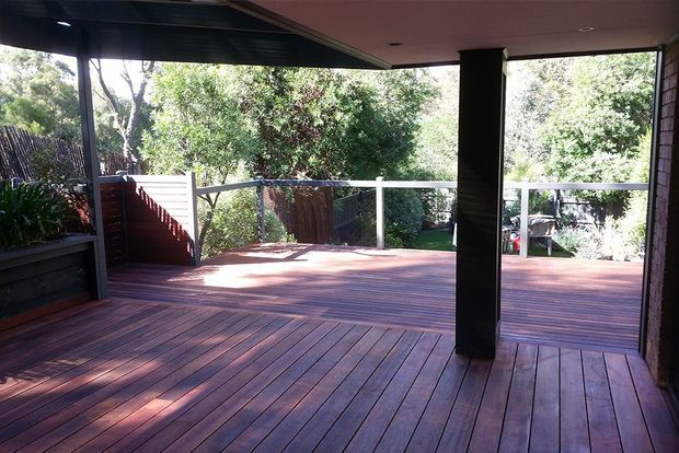Covered outdoor timber deck or terrace opening onto a leafy garden, with reddish-brown decking boards, dark structural posts and ceiling, glass or transparent balustrading, and built-in planter elements. The space appears connected to the house and is suitable for outdoor entertaining or relaxation.