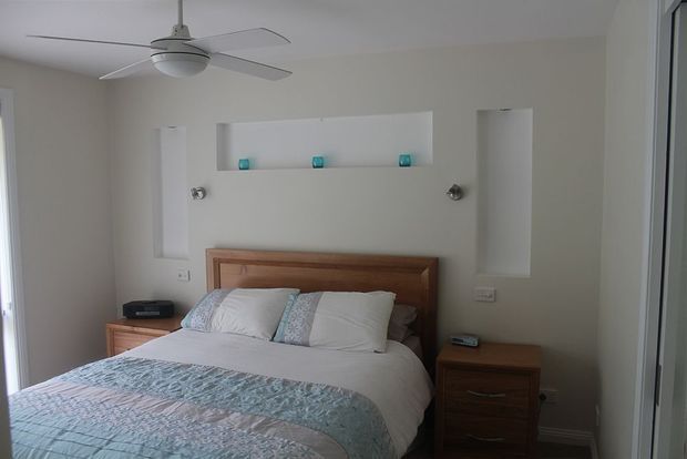 A calm, neutral-toned bedroom with a wooden bed frame and matching bedside tables, white bedding, and a pale blue patterned runner. The wall behind the bed features recessed display niches with small turquoise accents and wall-mounted reading lights. A white ceiling fan and built-in wardrobe or storage are visible.