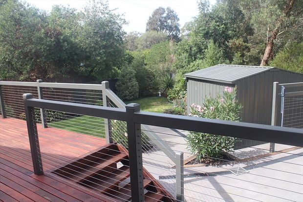 Elevated outdoor timber deck with dark cable-and-post balustrades, overlooking a paved courtyard and landscaped garden. A compact dark corrugated-metal-clad shed or outbuilding with a low-pitched roof sits among mature vegetation.