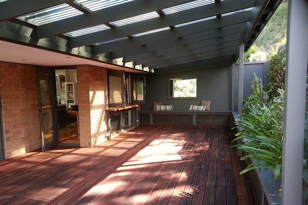 A covered outdoor deck or terrace attached to a brick house, with extensive timber decking, a built-in bench seat, dark structural beams, and translucent roof panels that provide filtered daylight. The space opens directly to the interior through glazed doors and is bordered by planted landscaping.