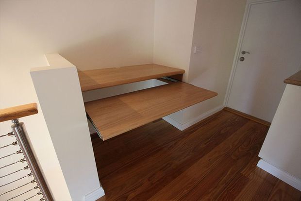 Compact interior stair landing with a custom timber staircase and integrated wooden bench-like treads extending from a white-painted wall structure. The space has dark timber flooring, white walls and trim, a white interior door, and stainless-steel cable balustrading visible at left.