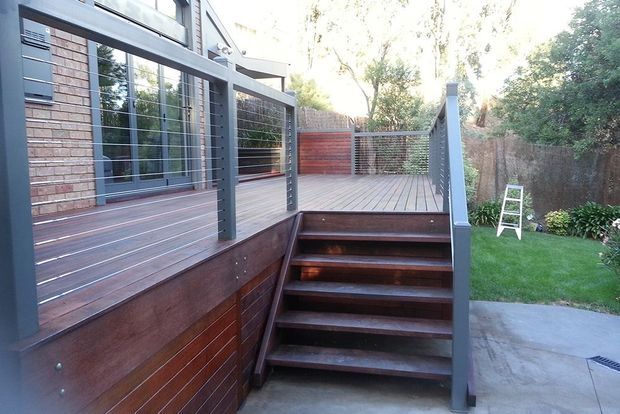 Exterior view of a contemporary timber deck and raised outdoor terrace adjoining a brick-and-clad house. Dark timber decking, stairs, privacy screening and grey metal cable-and-post balustrades frame a small landscaped backyard and paved area.