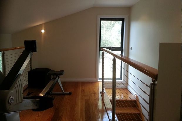 An upstairs landing or stairway with polished timber flooring, a timber handrail, and horizontal metal cable balustrades. A compact exercise machine sits beside the stairs, while a tall glazed door or window brings in natural light. The space has warm neutral walls and a simple contemporary residential fit-out.