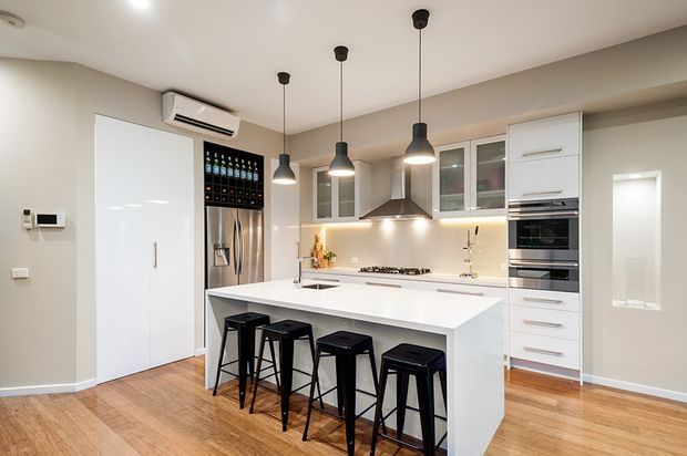 Bright contemporary kitchen with a large white island, four black bar stools, integrated stainless-steel appliances, white cabinetry, pendant lighting, and warm timber flooring. A built-in wine rack and refrigerator sit beside the island, with a cooktop and rangehood along the rear wall.