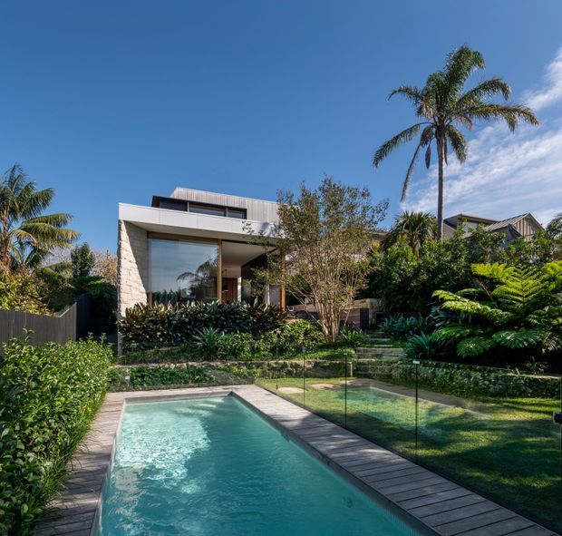 A contemporary residential exterior viewed from a landscaped backyard, featuring a narrow rectangular swimming pool, glass pool fencing, timber decking, dense tropical planting, and a box-like house with pale masonry walls, large glazing, and a low-pitched roof.