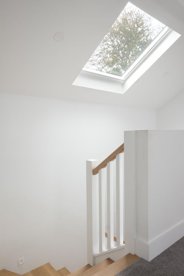 Bright minimalist stairway and landing with white painted walls, a timber handrail and stair treads, white vertical balustrades, grey carpet, and a large roof skylight providing natural light. The view looks down toward the staircase, with a partial upper-level wall and ceiling visible.