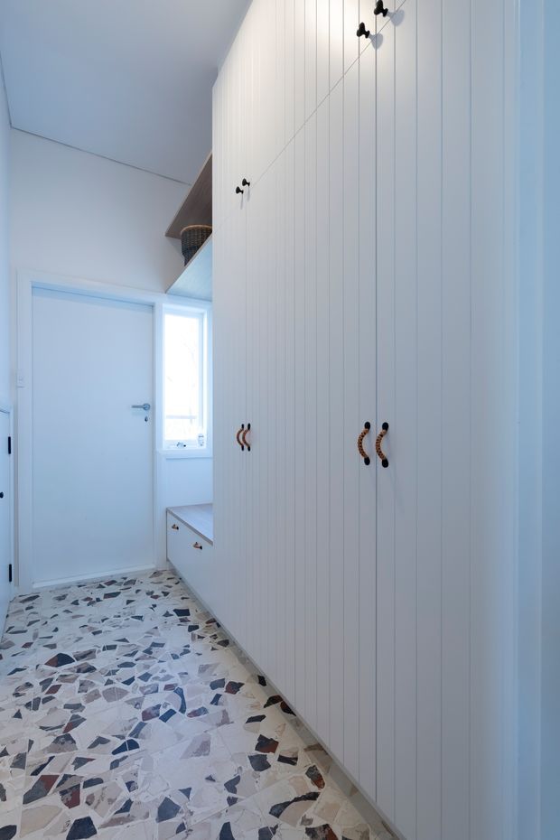 A narrow, bright hallway or entry storage area with full-height white vertical-panel cabinetry and dark curved handles. A white internal door and small window sit at the end, with built-in drawers below the window. The floor is finished in a distinctive multicoloured terrazzo-style surface with irregular grey, blue, beige and dark aggregate pieces. The space has a clean, light-filled contemporary feel.