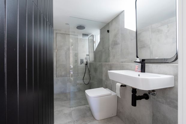 Contemporary compact bathroom with large grey stone-look wall and floor tiles, a frameless glass shower enclosure, wall-mounted white basin, concealed-cistern toilet, black tapware and exposed black vertical wall panelling. A rounded-rectangle mirror sits above the basin, creating a clean monochromatic scheme with strong black accents.