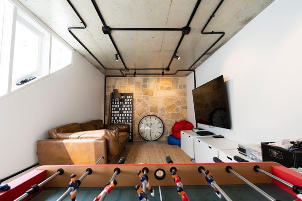 A compact contemporary media and games room with a foosball table in the foreground, leather sofas, a wall-mounted television, white low cabinetry, and a large analog clock. The room features an exposed concrete ceiling with black surface-mounted conduit and track lighting, a warm irregular stone feature wall, timber flooring, and high clerestory-style windows.