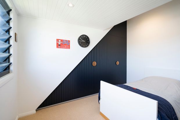 Bright bedroom with a white slatted ceiling, white walls, beige carpet, and a black vertically ribbed feature wall incorporating built-in storage doors. A bed with dark blue bedding occupies the foreground, with louvred window glazing on the left and simple wall decor and clock.