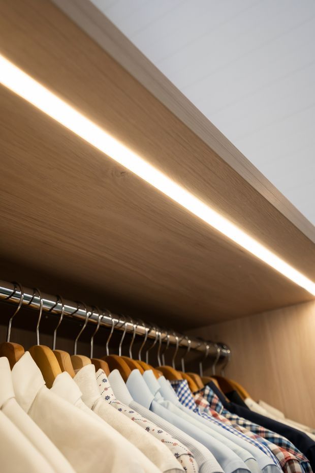 Close-up view inside a built-in wardrobe, showing a timber-finished hanging shelf with integrated linear LED lighting, a metal clothes rail, and neatly arranged shirts on wooden and metal hangers. The image emphasizes the custom wardrobe joinery and warm illumination.