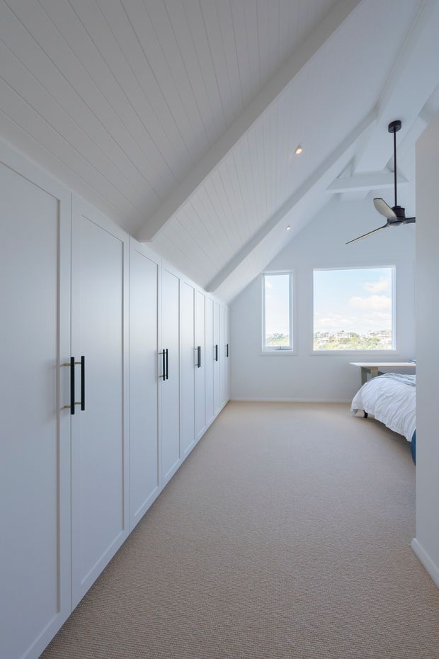 Bright, airy bedroom beneath a pitched, white-lined ceiling. The room features extensive built-in white wardrobe cabinetry with black linear handles along one wall, pale carpeted flooring, a partially visible bed, recessed lighting, a ceiling fan, and large windows overlooking a distant residential landscape.