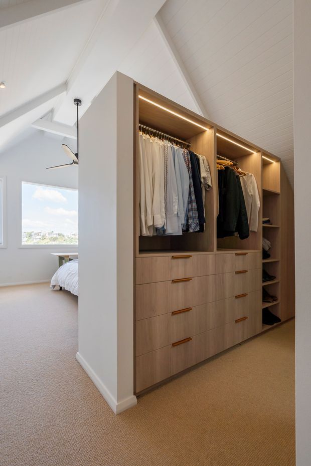 A bright built-in walk-in wardrobe beside a bedroom, featuring warm light-wood cabinetry with open hanging sections, drawers, shelving, and integrated strip lighting. The wardrobe sits beneath a white pitched, panelled ceiling, with beige carpet throughout. A bed, ceiling fan, and large window are visible in the adjoining bedroom.