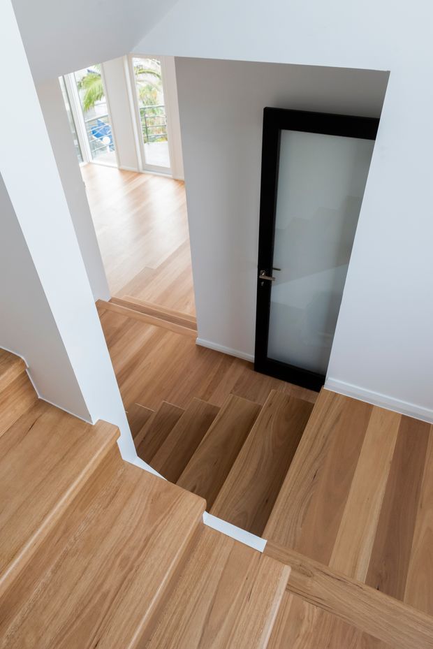 Bright contemporary stairway with warm timber flooring and stairs, white walls, and a black-framed frosted-glass door. The stairwell opens toward a light-filled landing with full-height glazed doors and views of greenery outside.