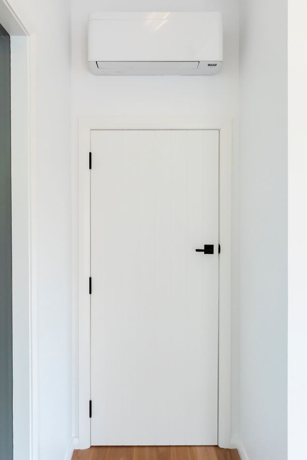 A minimalist interior hallway or entry area with a tall white flush-panel door, black hinges and lever handle, white walls and trim, and warm timber flooring. A white split-system air-conditioning unit is mounted above the door. The scene is bright, clean, and sparsely detailed.