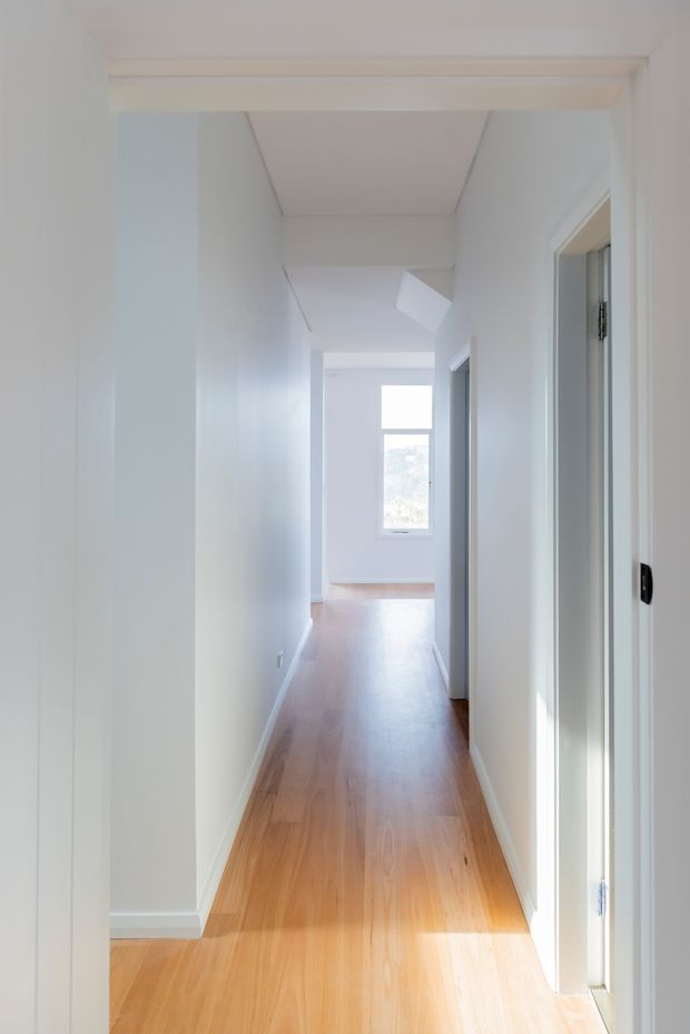 A bright, narrow residential hallway with white walls, white trim and doors, warm timber flooring, and natural light entering from a window at the far end. A small skylight or ceiling opening adds additional daylight, creating a clean and airy passage.