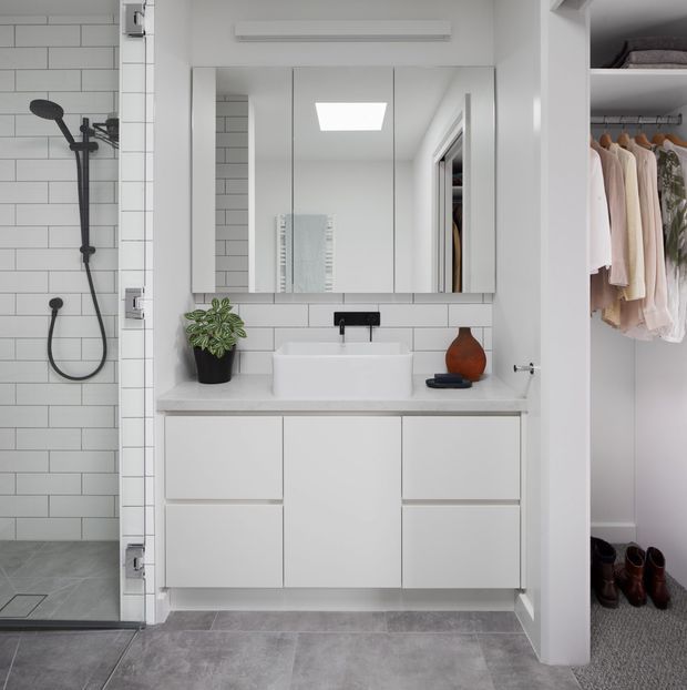Bright, compact modern bathroom with a walk-in tiled shower, wall-mounted white vanity, rectangular basin, mirrored shaving cabinet and black tapware. An open walk-in wardrobe is visible directly beside the bathroom, with hanging clothes and shoes. The room uses white subway wall tiles, grey floor tiles and minimalist built-in joinery, with a small potted plant and decorative vessel adding warmth.