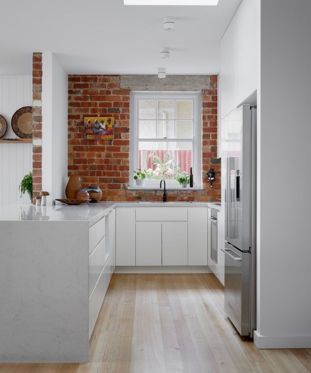 Bright contemporary kitchen with white cabinetry and a large white stone island, set against exposed red brick walls and a rendered ceiling. The room includes stainless-steel appliances, a timber floor, a double-hung window, integrated lighting, and restrained decorative styling.