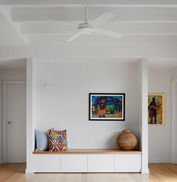 Bright, minimalist built-in display and storage wall in a white interior. The feature has vertical white wall panelling, a warm timber bench top, and white base cabinetry with flat fronts. A framed colourful artwork, woven decorative vessel, and patterned cushions add visual interest. Pale timber flooring, white doors, and a white ceiling fan create a clean, airy residential setting.