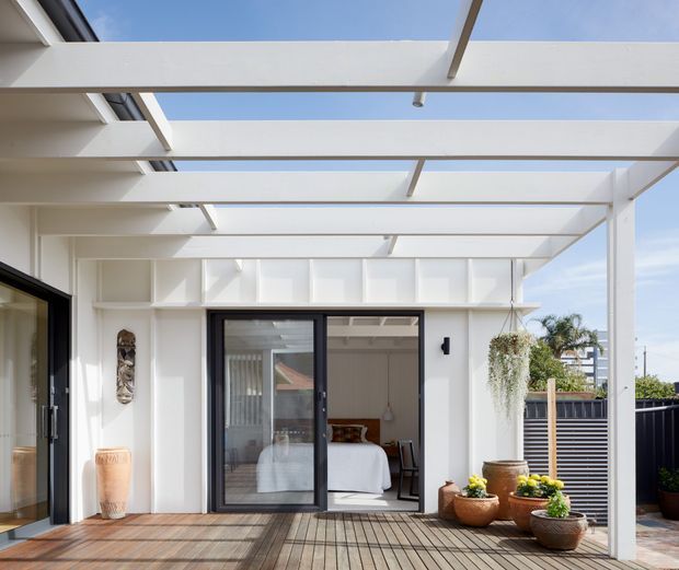 Bright contemporary outdoor deck beneath a white pergola with open skylight-like roof panels and exposed beams. Black-framed glazed doors open into a bedroom, while potted plants, timber decking, and a slatted privacy fence define the terrace. The image emphasizes the indoor-outdoor connection and airy architectural structure.