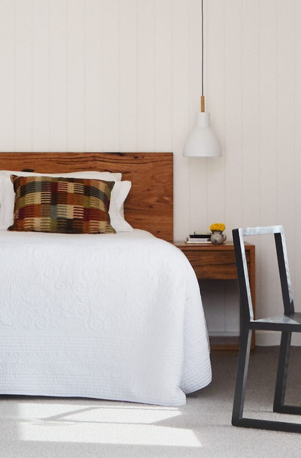 A bright, calm bedroom featuring a white textured bedspread, a substantial natural timber headboard and bedside table, white vertical wall panelling, a suspended white pendant light, and a dark framed chair. A patterned multicolour accent cushion adds warmth against the predominantly neutral scheme.