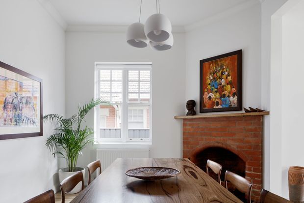 Bright dining room with a substantial live-edge timber dining table, leather and timber dining chairs, a red brick open fireplace, white walls and ceiling, plantation shutters, indoor palms, framed artwork, and sculptural pendant lighting.