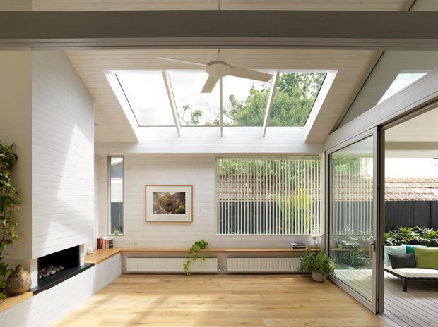 A bright, contemporary sunroom or living area with a glazed lean-to skylight and central ceiling fan. The room features white-painted brick walls, light timber flooring and ceiling boards, built-in low timber cabinetry and shelving, a white brick fireplace with dark hearth, and large sliding glass doors opening to an outdoor deck. Vertical screening, garden planting, and outdoor seating are visible beyond the glazing.