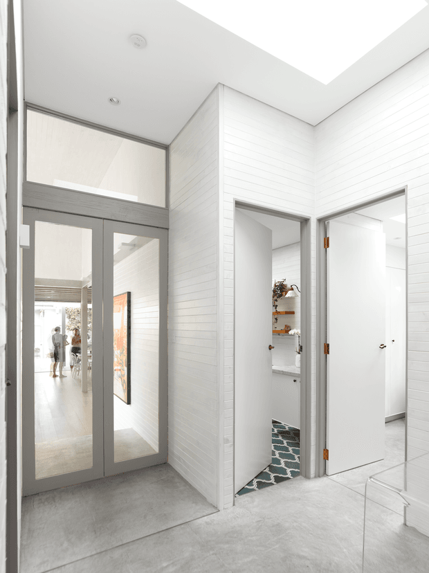 Bright contemporary hallway with white horizontal wall panelling, pale grey concrete-look flooring, and white doors with grey trim. A full-height glazed internal door opens toward a light-filled living area, while an open doorway reveals a compact bathroom with patterned floor tiles and timber shelving. A large rectangular skylight brings in natural light, creating an airy, minimal interior.