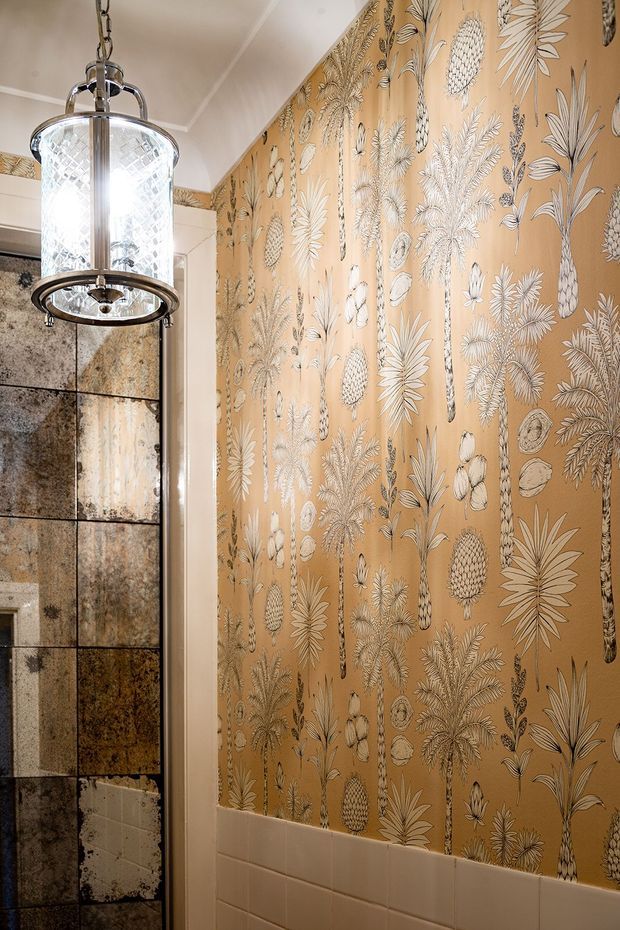 A close-up view of a richly decorated bathroom featuring a warm tan botanical and pineapple-patterned wallpapered wall above white rectangular wall tiles. A vintage-style chrome pendant light with textured glass hangs in the foreground, while the adjacent wall is clad in varied dark, distressed stone-look tiles. The space has an ornate, eclectic and warm character.