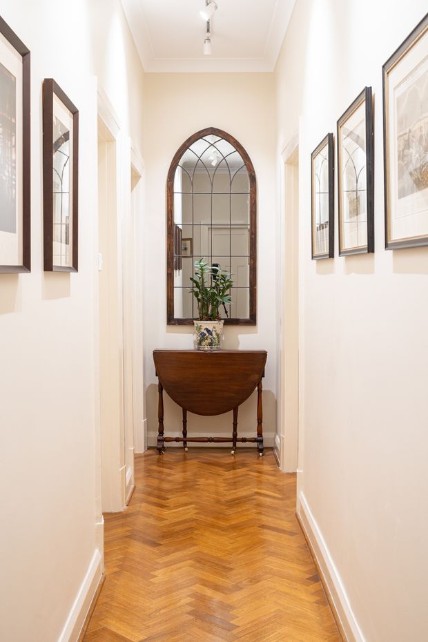 A narrow, bright residential hallway with cream painted walls, white trim, framed artwork, and a polished honey-toned herringbone timber floor. At the far end is a traditional dark timber console table beneath a tall arched leaded-glass mirror, with a potted plant adding greenery. Ceiling-mounted spotlights illuminate the gallery-like corridor.