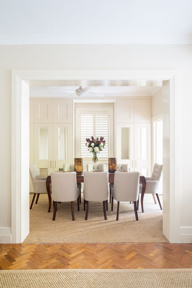Bright, refined dining room viewed through a wide white-trimmed opening. A long dark wood dining table is surrounded by upholstered dining chairs, with a floral arrangement and decorative candleholders at the centre. The room features cream built-in cabinetry with inset panels and mirrored doors, plantation shutters, a white ceiling fan, and a textured natural-fibre rug. The foreground has warm herringbone timber flooring and a neutral woven runner.