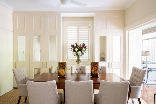 Bright, elegant dining room with a large dark timber dining table surrounded by upholstered dining chairs. The room features extensive cream built-in cabinetry with paneled doors and integrated mirrored panels, a central window with white plantation shutters, pale walls, timber flooring, decorative table lamps, and a floral arrangement. A ceiling fan and adjoining bright room are visible through the opening at right.