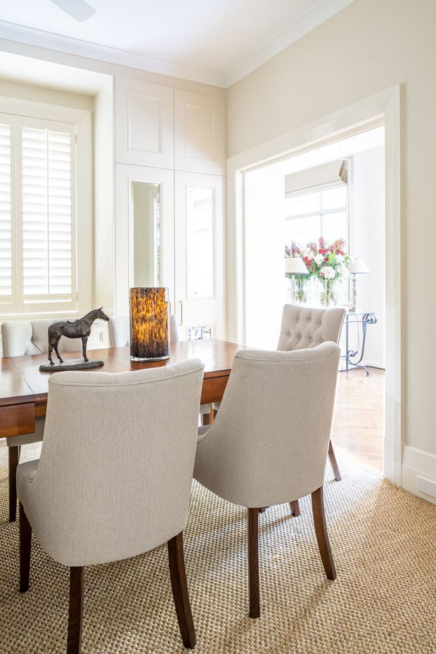 Bright, elegant dining room with a rectangular wooden dining table surrounded by upholstered high-back chairs. The room features white plantation shutters, detailed white built-in wall panelling and cabinetry, broad traditional architraves, pale neutral walls, a woven natural-fibre rug, and a large opening into a light-filled adjoining sitting area. A glass vase and decorative objects sit on the table.