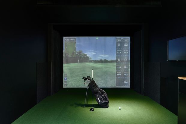 A dark, enclosed indoor golf simulator room with a large projection screen showing a virtual golf course, a golf bag and clubs on artificial turf, and surrounding black acoustic wall panels. A small illuminated display or screen is visible on the right wall.