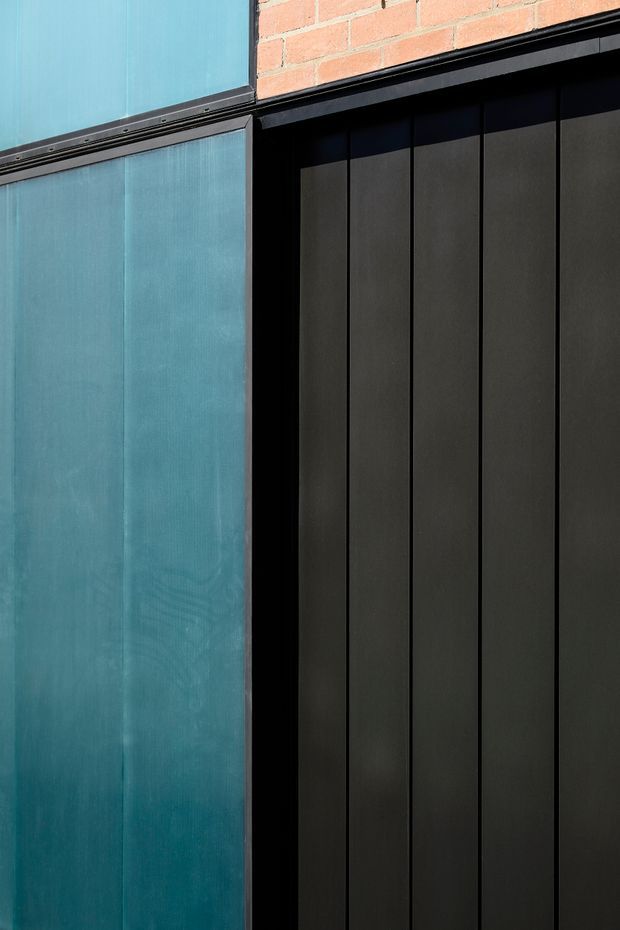 Close-up architectural detail showing contrasting vertical facade panels: a blue-green translucent or textured panel system on the left and dark charcoal-black narrow-panel cladding on the right, framed with black metal trims beneath a red brick wall.