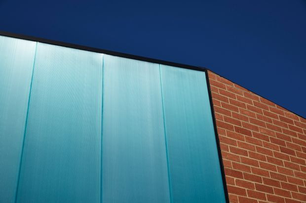 Close-up exterior architectural detail showing a large translucent blue-green corrugated or ribbed panel framed in dark metal beside a red-orange brick wall. The sharply angled roofline and deep blue sky create a bold, high-contrast composition.