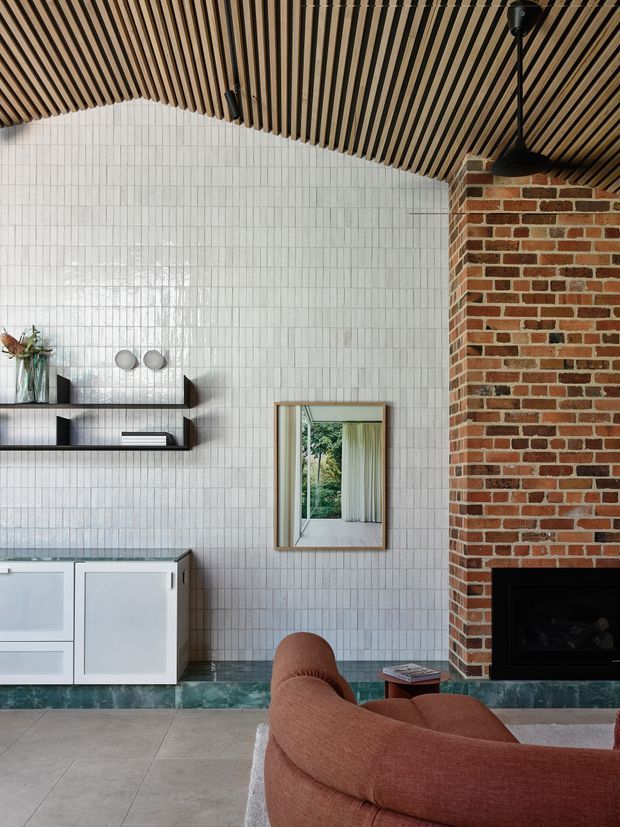 A refined living room with a dramatic angled timber slatted ceiling, white small-format tiled walls, a red exposed-brick fireplace, dark green stone skirting, and pale stone flooring. Built-in white cabinetry and minimalist black shelving line the wall, while a curved rust-coloured sofa occupies the foreground. A framed mirror reflects greenery and a curtained window, adding depth to the room.