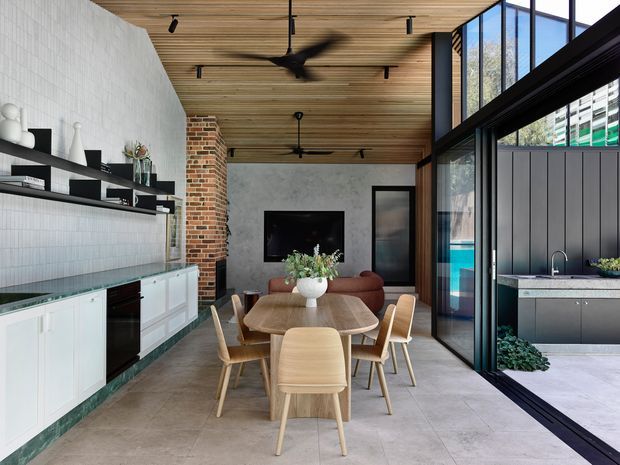 Open-plan dining and kitchen interior with a timber-lined ceiling, pale tiled flooring, white tiled and painted kitchen joinery, exposed brickwork, and black-framed full-height glazing. A timber dining table with light timber chairs sits beside a compact living area, while the glazing opens visually toward an outdoor kitchen or courtyard area.
