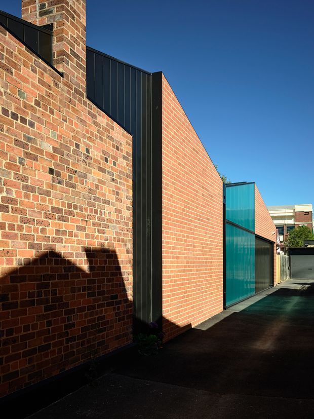 Exterior architectural view of a narrow urban passage beside a contemporary brick building. The facade combines warm red and multicoloured face brickwork with tall black vertical metal cladding elements and a striking turquoise translucent glazed wall. Dark paving and strong sunlight create sharp shadows, with neighbouring urban buildings visible in the distance.